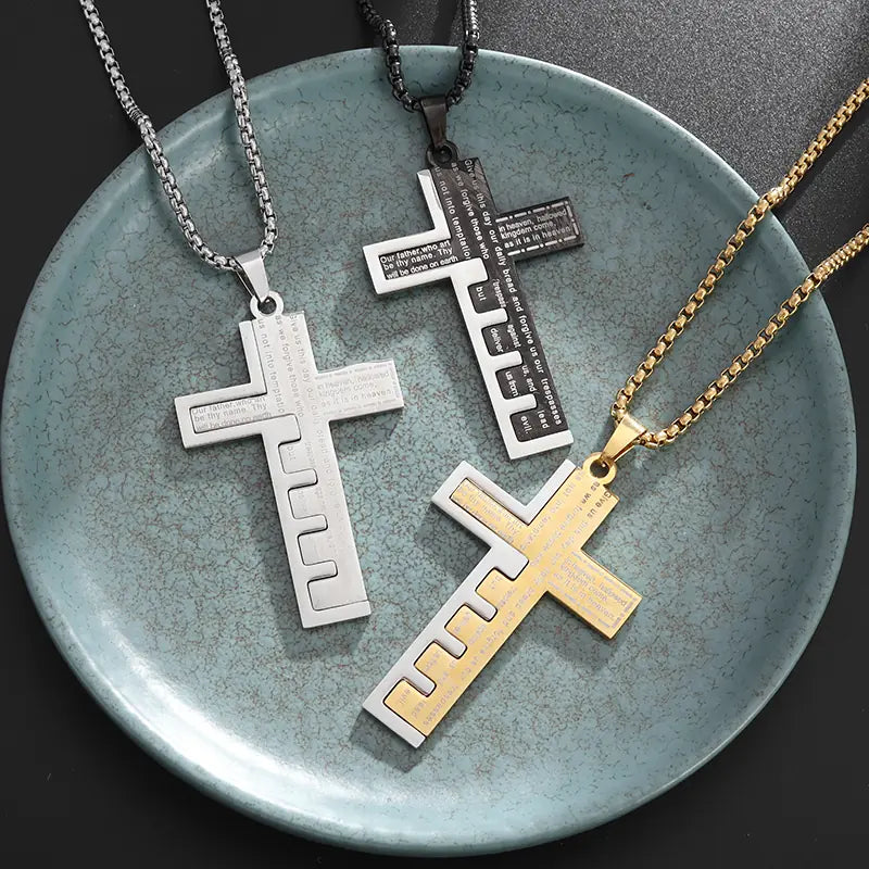 Colección Escudo Divino con tres dijes de la Cruz del Padre Nuestro alineados en variantes de color plata clásica, negro mate y oro dorado sobre un plato cerámico. Accesorios religiosos de alta gama y joyería de acero quirúrgico para hombres en México.