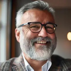 Retrato de un hombre elegante de aproximadamente 60 años, con cabello y barba canosos, usando gafas de armazón negro y saco de vestir. Testimonio de cliente satisfecho de la Cruz del Padre Nuestro, collar para hombre en acero inoxidable que refleja fe, elegancia y fortaleza. Joyería masculina premium para caballeros.
